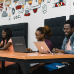 A group of young Black professionals work together in a modern co-working space, seated along a long wooden table with laptops open. how to start an AI automation agency in Nigeria