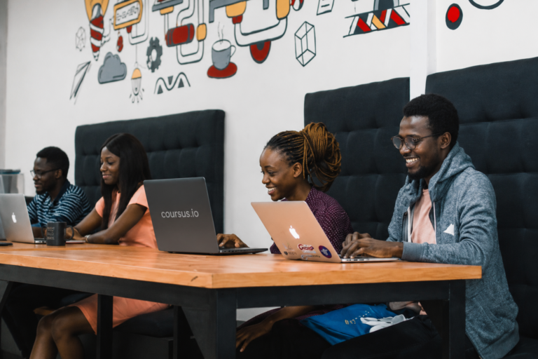 A group of young Black professionals work together in a modern co-working space, seated along a long wooden table with laptops open. how to start an AI automation agency in Nigeria
